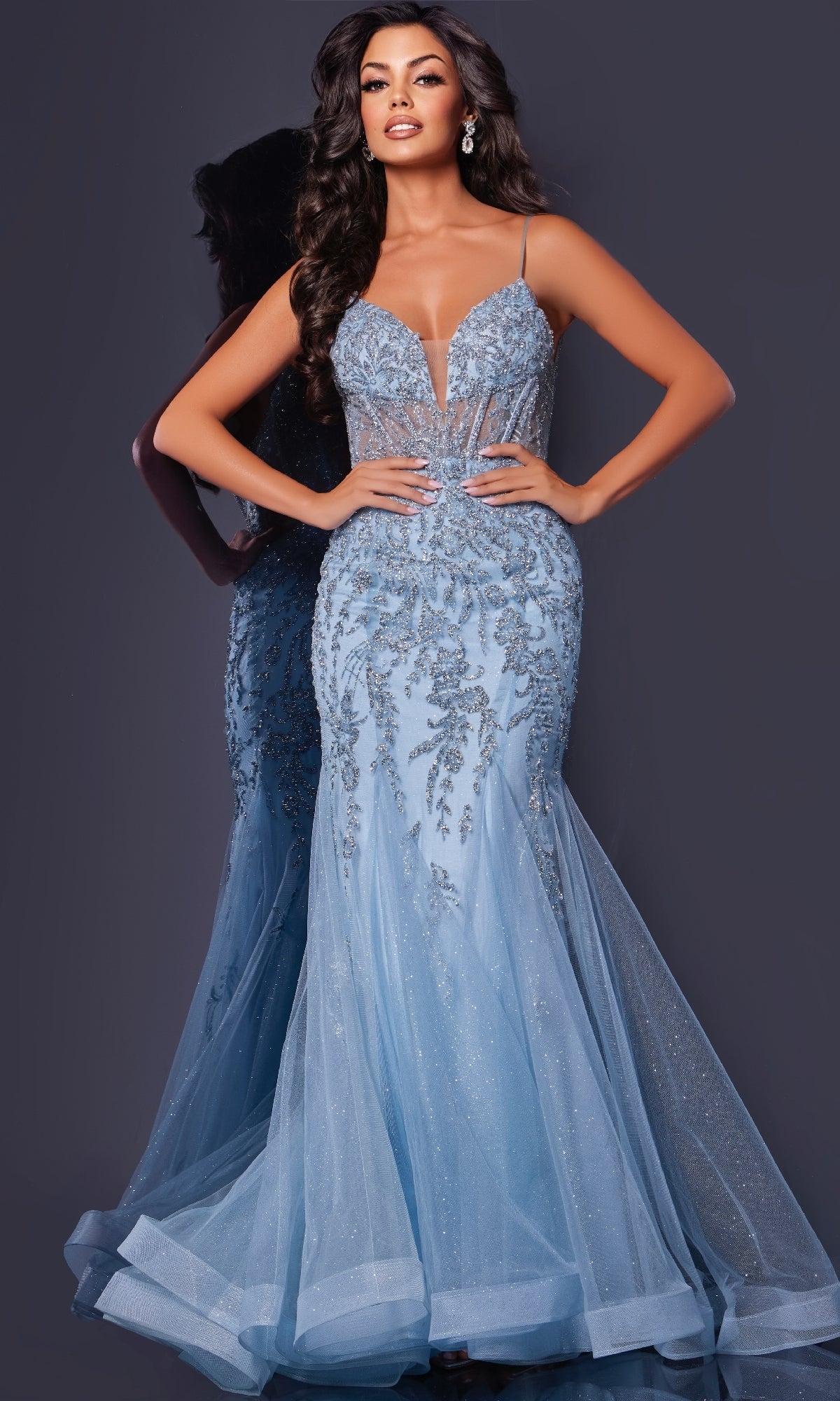A woman wears the Jovani 43845 long prom dress—a fitted, sparkly light blue gown with floral sequin embroidery, thin straps, and a sheer tulle skirt—while standing against a dark background.